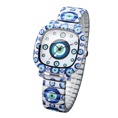 A square analog watch with a white face and a matching metal bracelet, both decorated with a blue and green evil eye pattern. The watch is photographed on a dark textured surface with a white box edge visible in the background.